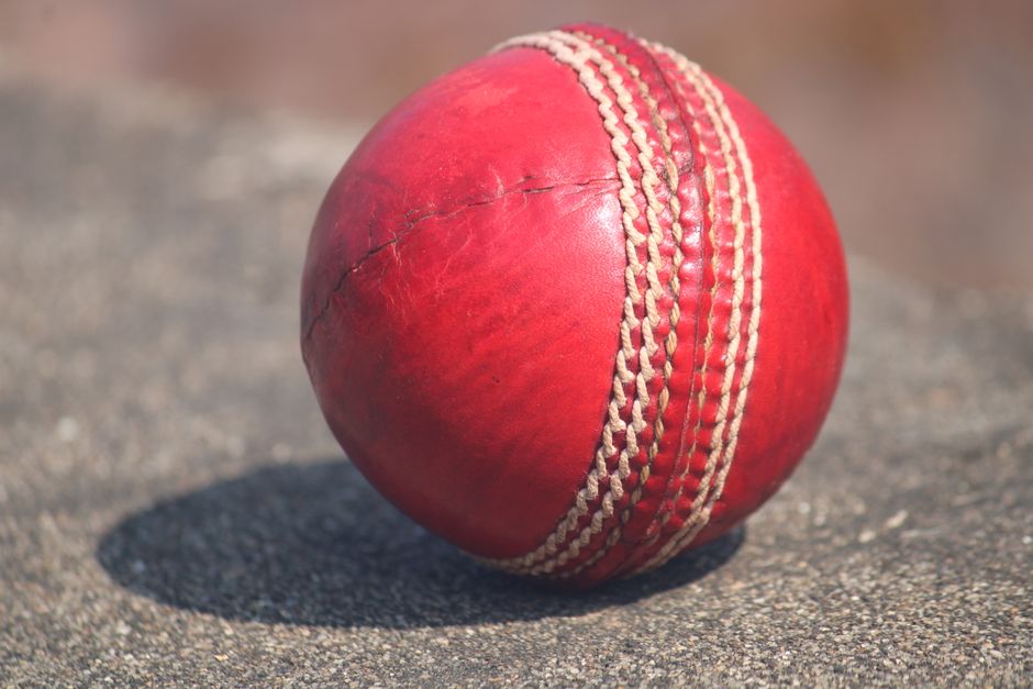 A close-up photograph of a well-worn red cricket ball resting on a textured asphalt surface, casting a distinct shadow beneath it. The ball features visible scuffs and scratches, with a prominent whit